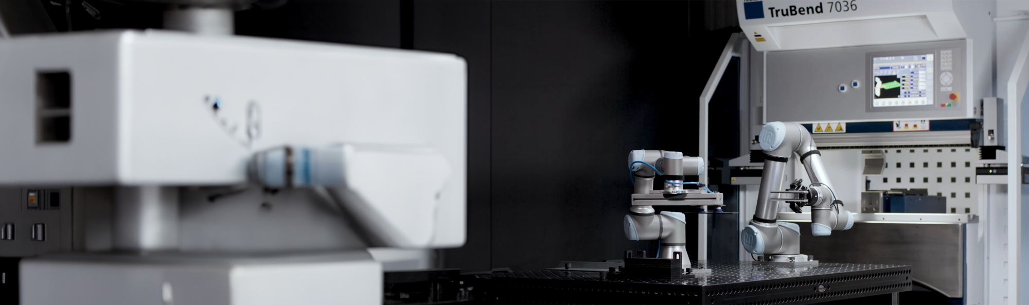 Das Bild zeigt einen modernen Arbeitsbereich mit Industrierobotern, im Vordergrund ist ein Roboterarm unscharf zu sehen, während im Hintergrund zwei weitere Roboterarme bei der Bedienung einer Maschine, die als "TruBend 7036" gekennzeichnet ist, fokussiert abgebildet sind.