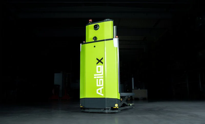  Das Bild zeigt einen grünen, automatisierten Fördertechnik-Roboter mit der Aufschrift "Agilox", der für den industriellen Einsatz konzipiert ist.