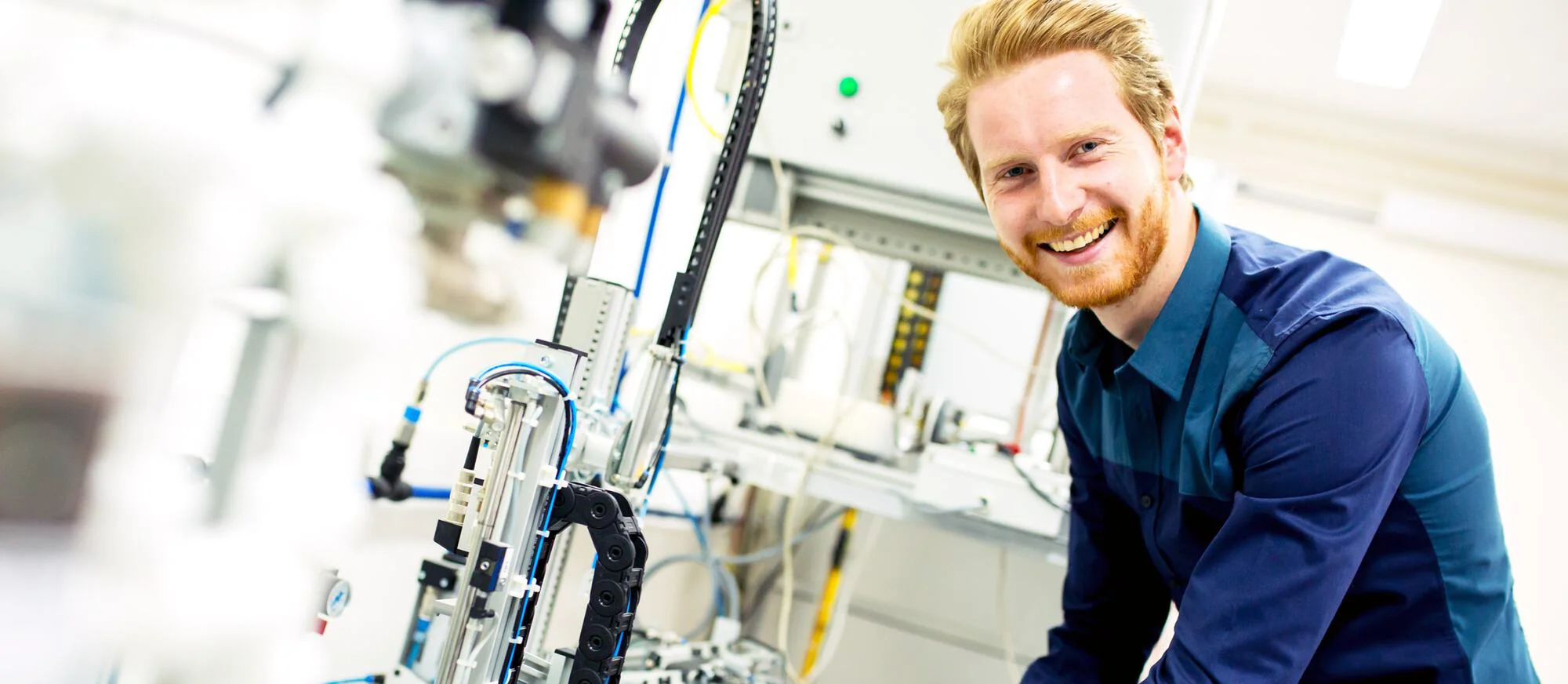 Das Bild zeigt einen lächelnden Mann mit rotem Haar und Bart, der ein blaues Hemd trägt und neben einer komplexen technischen Ausrüstung oder einem Roboter in einer hellen Laborumgebung steht.