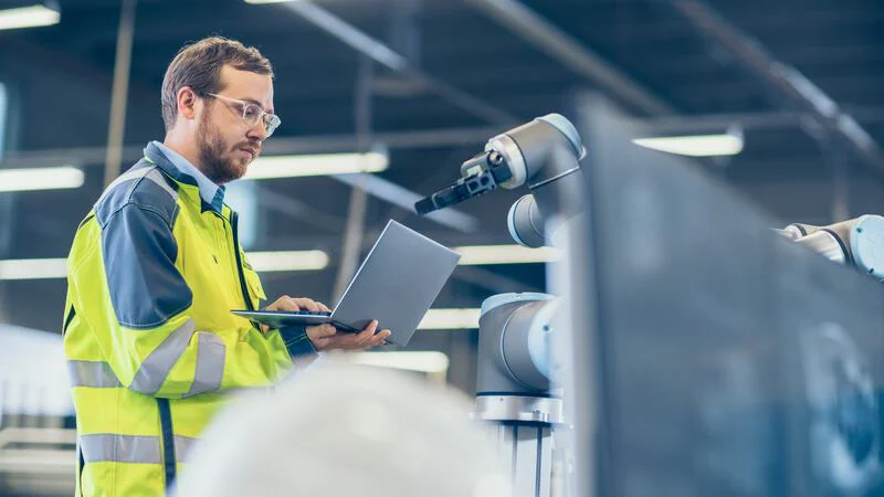 Das Bild zeigt einen fokussierten Arbeiter in einer Sicherheitsweste, der ein elektronisches Gerät bedient, neben einem industriellen Roboterarm in einer Werkstattumgebung.
