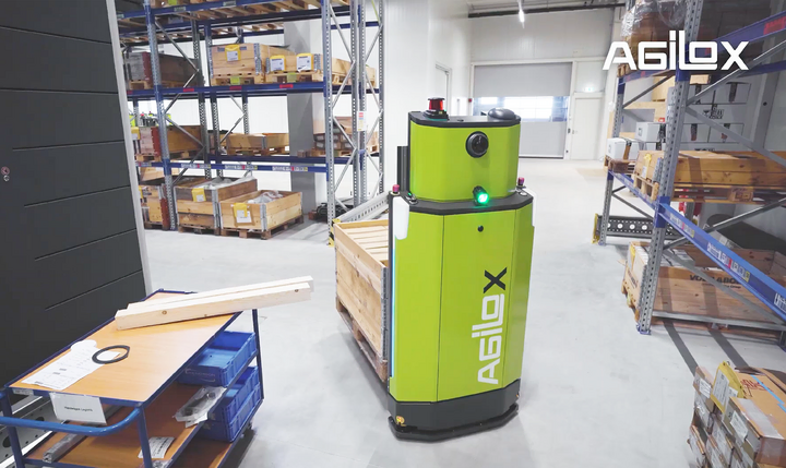 Auf dem Bild ist ein leuchtend grüner, zylindrischer AGILOX Autonomer Mobiler Roboter zu sehen, der in einem Lager mit Regalen und Lagergut im Hintergrund navigiert.